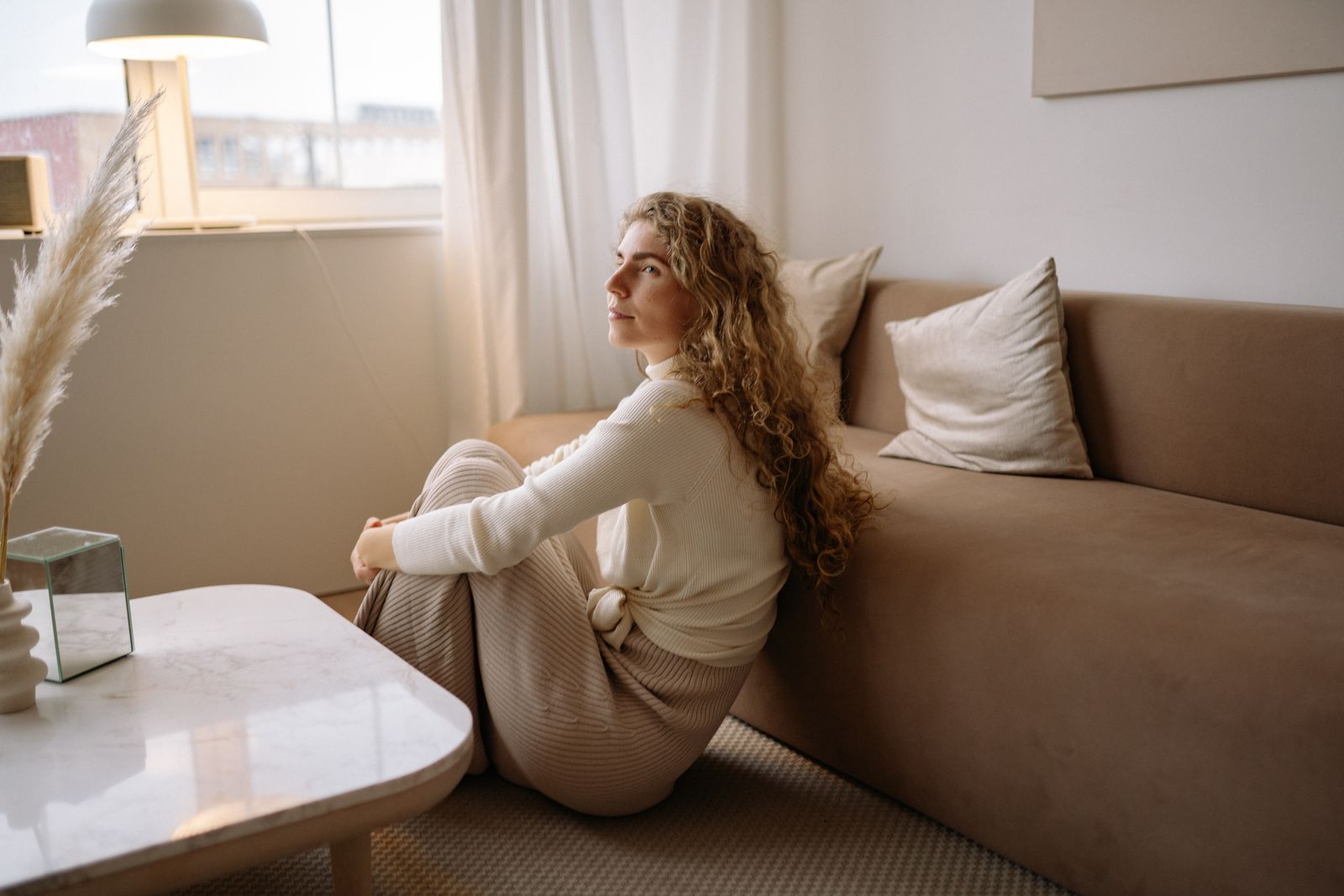 Mujer sentada en el suelo apoyada en el sofá, con la mirada hacia la ventana, en un salón de tonos cálidos